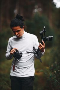 A male photographer in a gray sweater holding a camera and gimbal outdoors, surrounded by greenery.